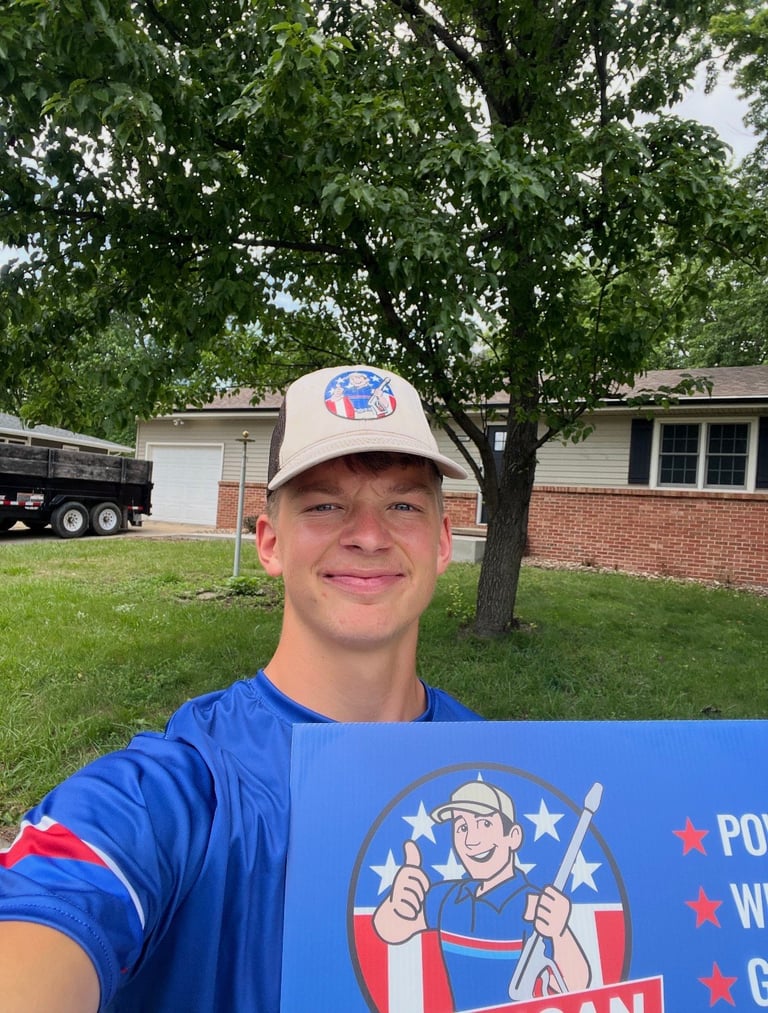 Drew in a blue American Softwash shirt and tan-and-mesh cap, smiling in front of a brick home in Des Moines, Iowa