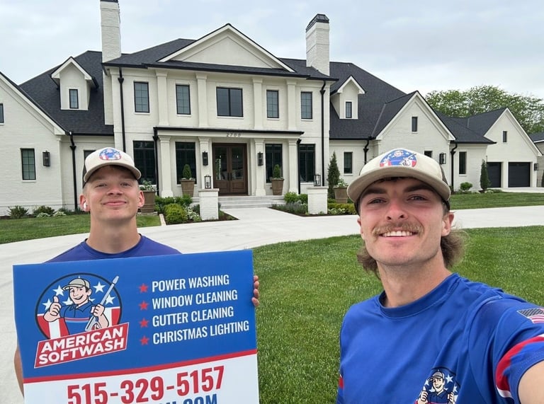 Drew and a coworker in matching American Softwash shirts holding the company sign in front of a large white home in Des Moines