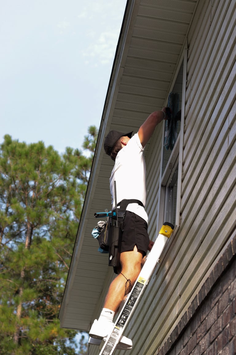 Worker in white shirt and black shorts installing a solar panel on a metal roof with a ladder leaning against the building