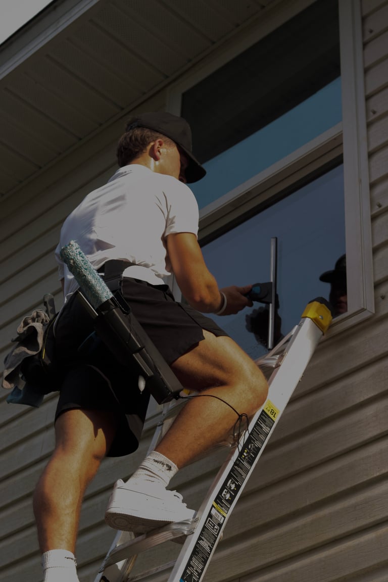 Worker climbing a ladder while installing a window or vent on a beige-sided residential home with tools on his belt