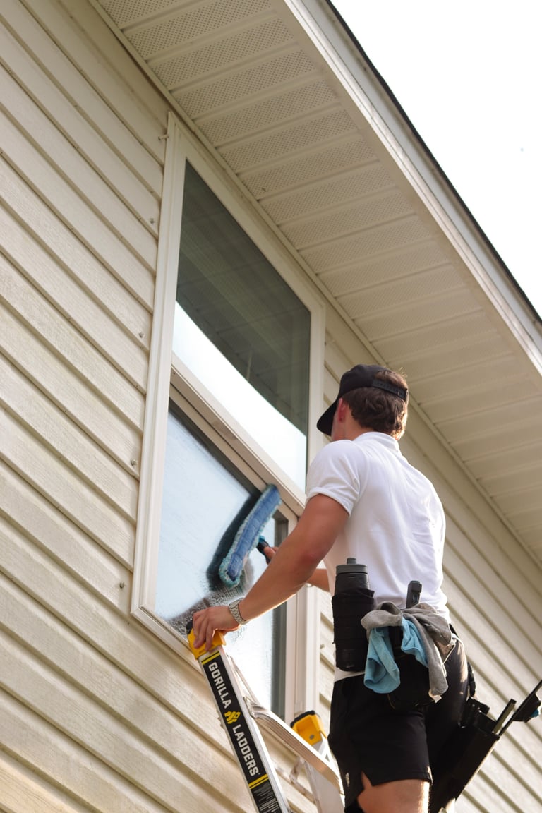 Worker in white shirt and black cap squeegeeing a large residential picture window from a Gorilla ladder, with cleaning supplies in a tool belt