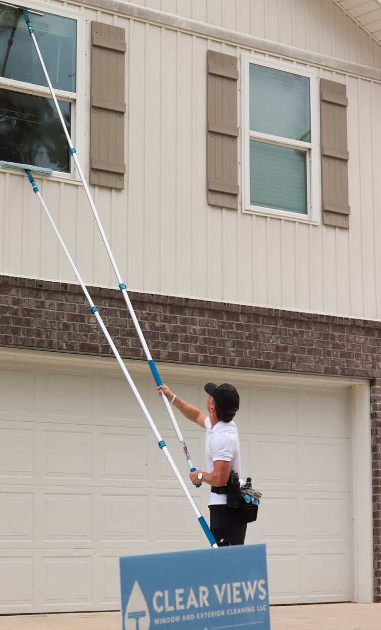 Drew on an extension pole cleaning the second-story windows of a brick and siding Pensacola home, with the blue Clear Views Window and Exterior Cleaning LLC yard sign set out in front