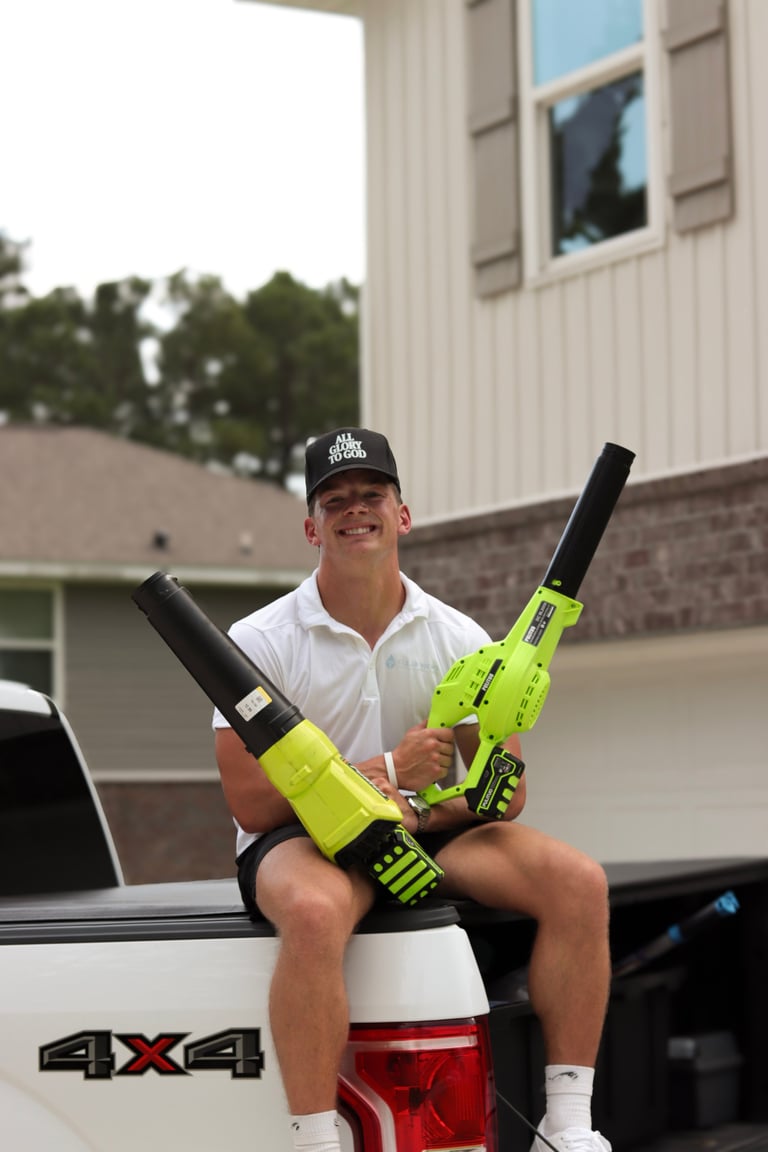Man in white shirt and black cap sitting on white pickup truck bed holding two neon yellow nail guns