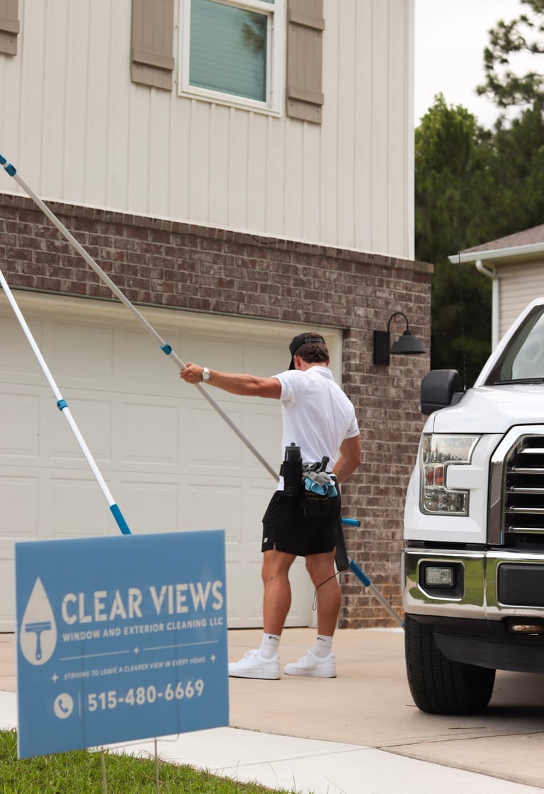 Worker in white shirt and black cap cleaning residential windows with extended pole while white pickup truck is parked in driveway