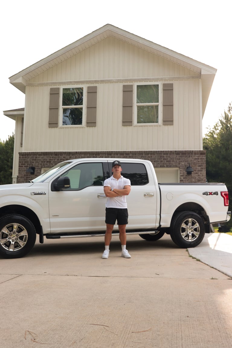 Man standing in front of a white pickup truck parked in the driveway of a two-story brick and siding house