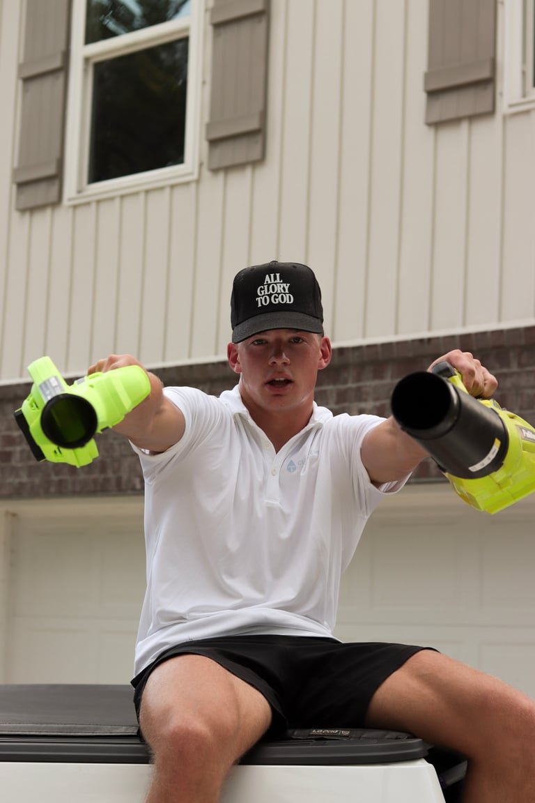 Man in white shirt and black cap wearing neon safety vests, holding construction pipes while sitting outdoors against a white building