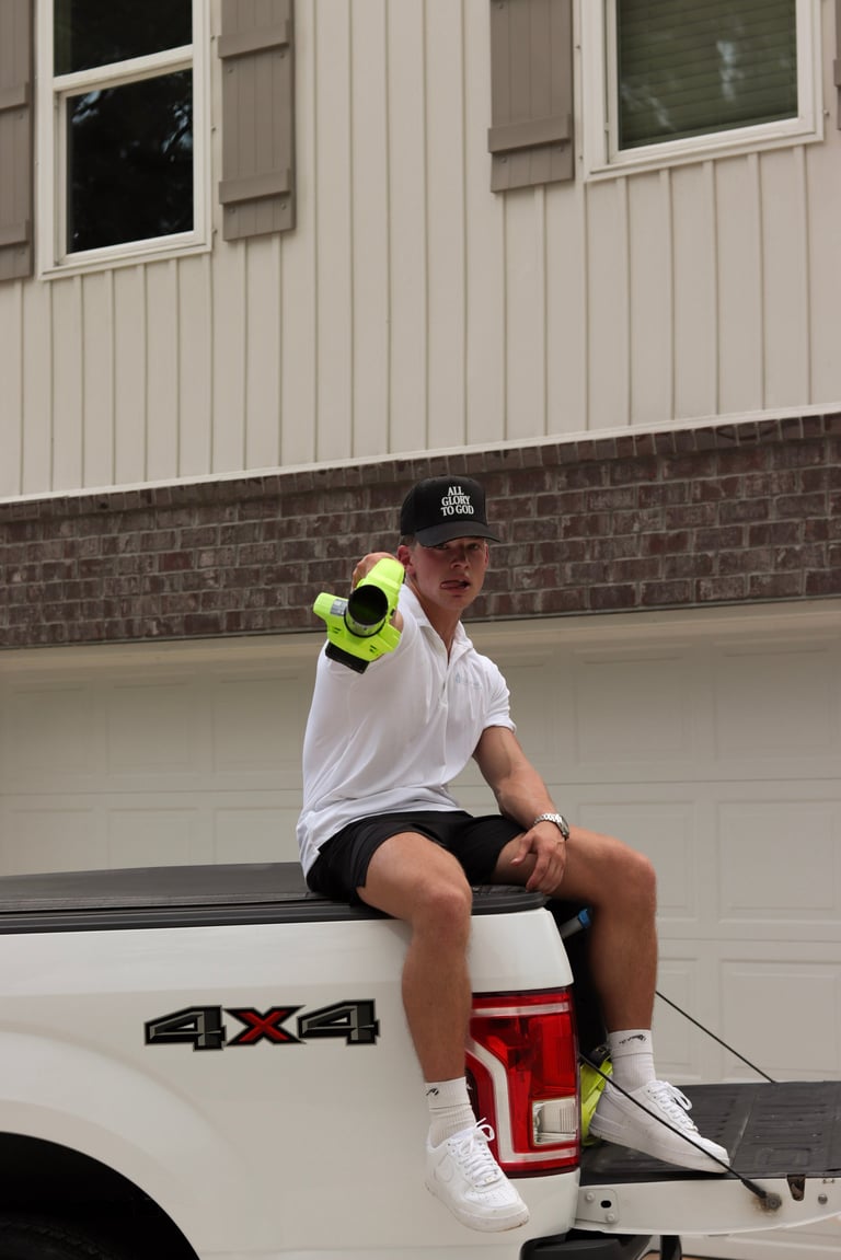 Drew sitting on the tailgate of the white 4x4 Clear Views pickup truck, holding a neon yellow binoculars and aiming it forward