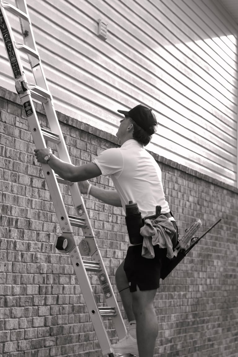Man in white shirt climbing an aluminum ladder against a brick wall under a corrugated metal overhang