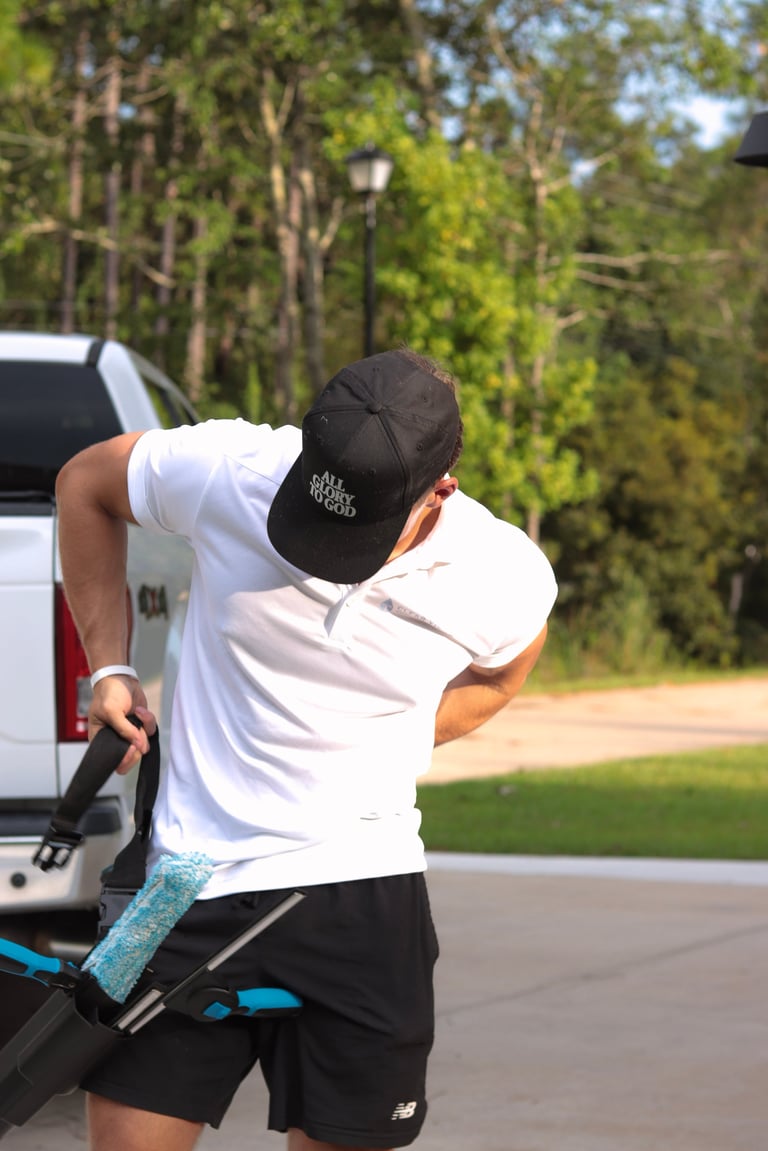 Man in white shirt and black cap adjusting a backpack near the white company truck parked on a residential driveway with trees in the background