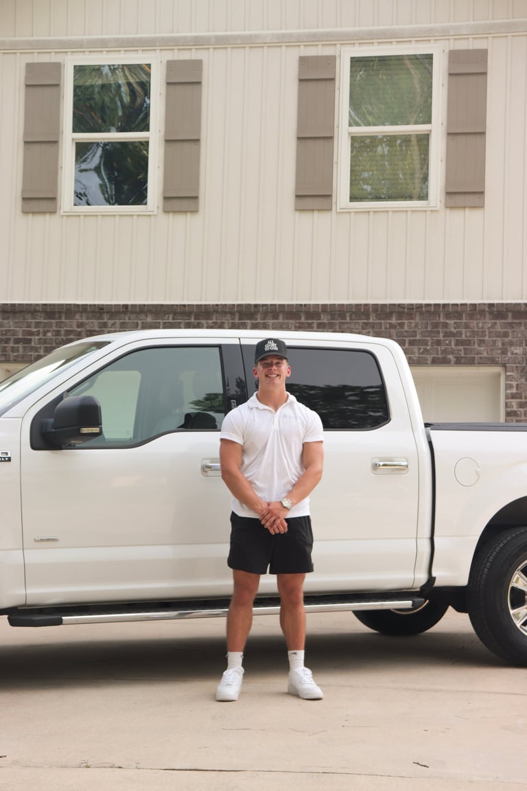 Drew in a white polo and black 'All Glory to God' cap standing in front of the white Clear Views pickup truck parked in a Pensacola driveway