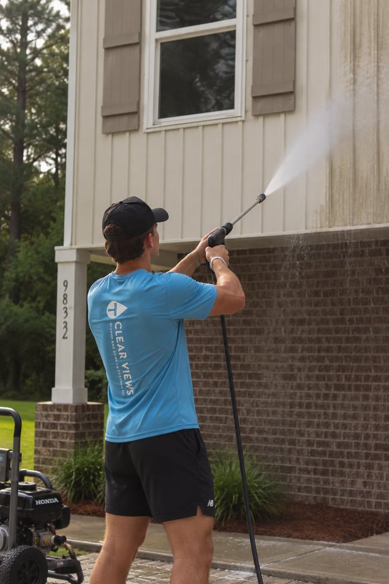Worker in a blue Clear Views t-shirt spraying water on the brick and siding exterior of a house with a pressure washer, visible Honda-powered unit in the foreground