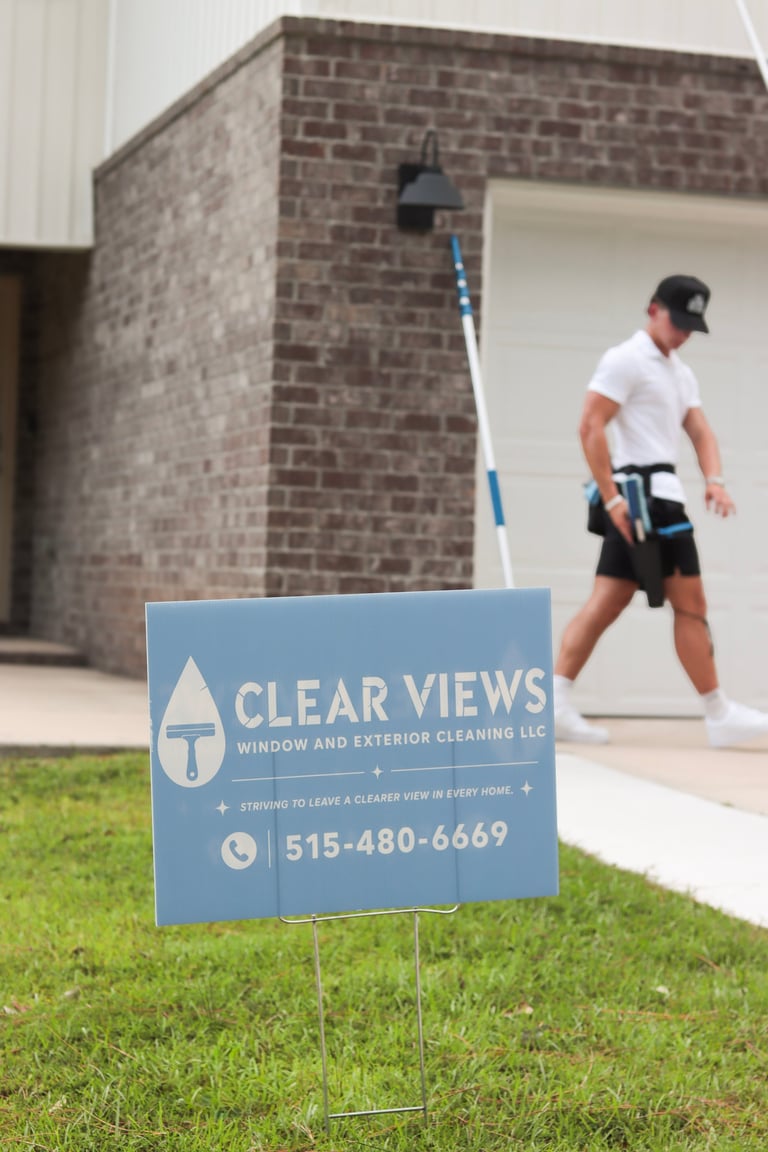 Worker with long-handled brush cleaning house exterior next to Clear Views Window and Exterior Cleaning LLC yard sign