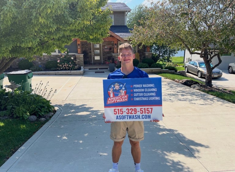Drew standing on a Des Moines residential driveway holding the blue American Softwash sign advertising power washing, window cleaning, gutter cleaning, and Christmas lighting