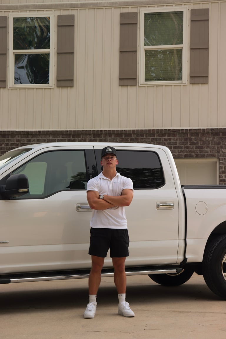 Man in white shirt and black shorts standing with arms crossed in front of the white pickup truck against a brick and metal building