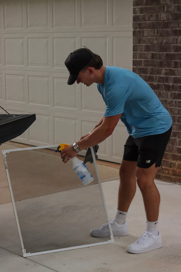 Man in blue shirt and cap using a power drill on a screen frame resting in the driveway in front of white garage door