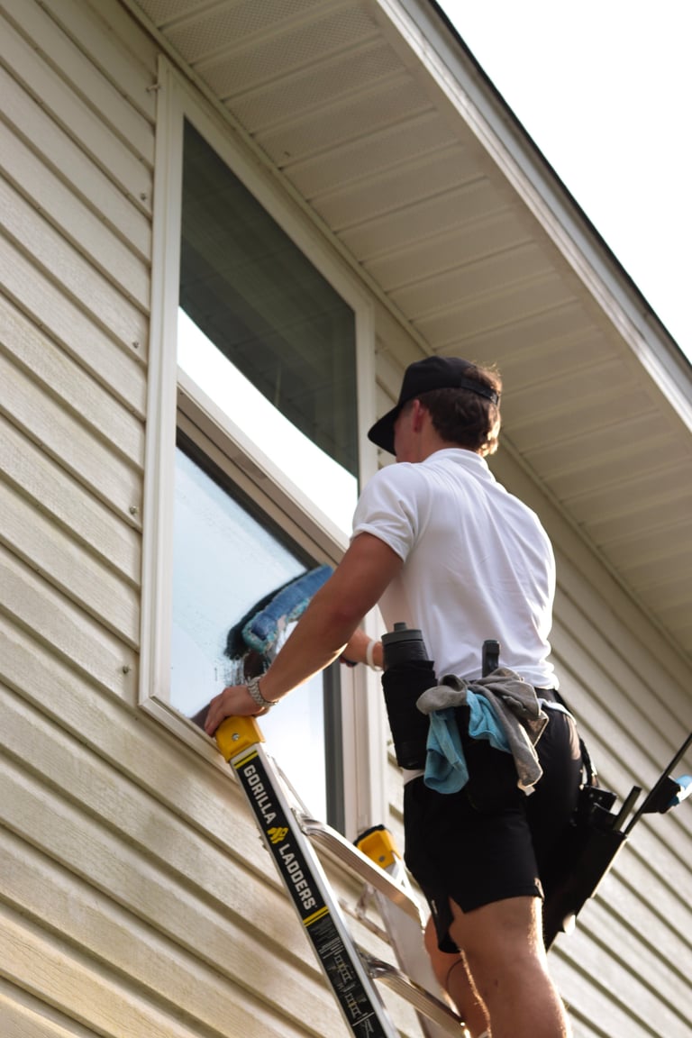 Worker on a Gorilla ladder installing or repairing a window on a beige vinyl-sided house exterior