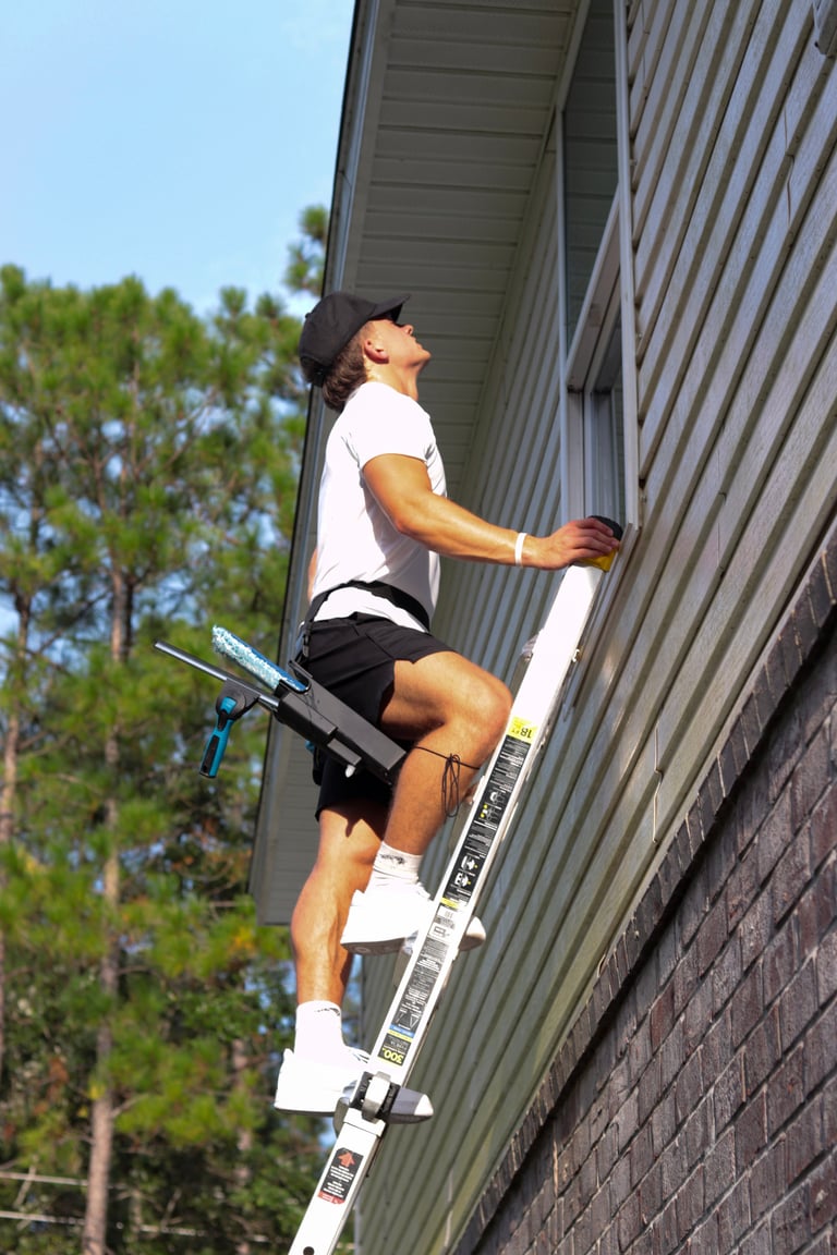 Drew climbing a tall white extension ladder against a corrugated metal building while working a window with his squeegee and tools on his belt