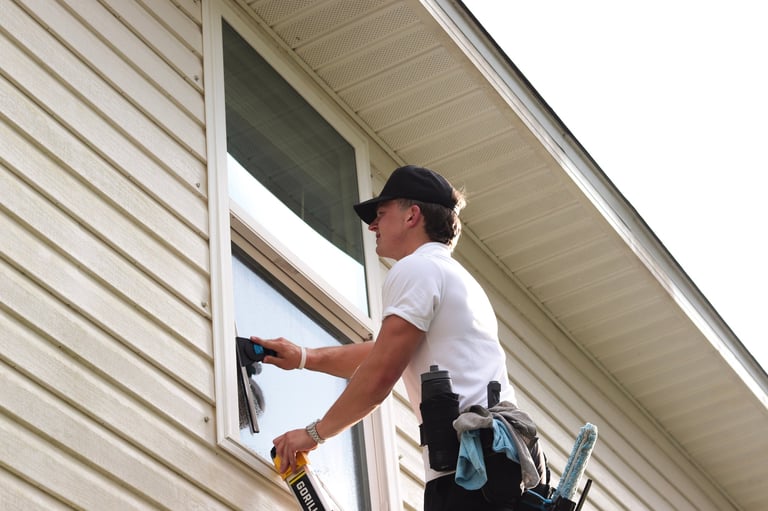 Worker cleaning an upper window on beige vinyl siding while standing on a ladder with squeegee and cleaning tools